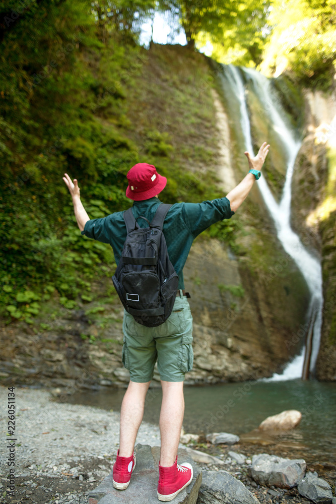 Young male caucasian traveler in red hat and summer hiking outfit enjoying  a waterfall during his single trip at the wooden mountains, back view.  Photos