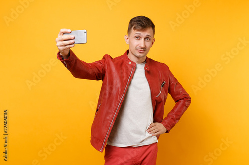 Portrait vogue handsome young man in red leather jacket, t-shirt doing selfie on mobile phone isolated on bright trending yellow background. People sincere emotions lifestyle concept. Advertising area © ViDi Studio