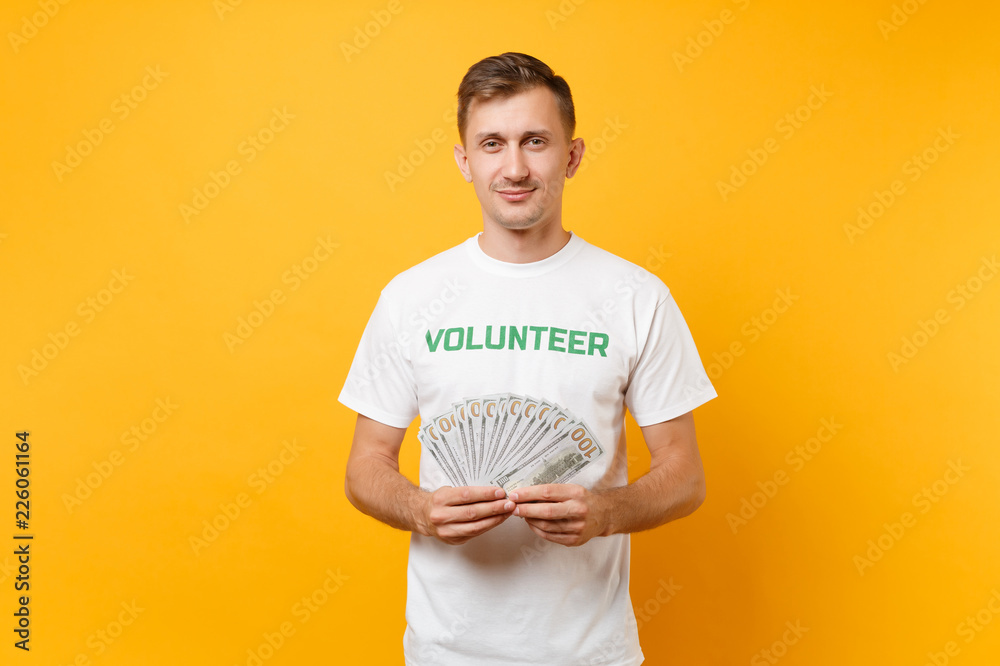Man in white t-shirt with written inscription green title volunteer hold lots dollars banknotes, cash money isolated on yellow background. Voluntary free assistance help, charity grace work concept. Man in white t-shirt with written inscription green title volunteer hold lots dollars banknotes, cash money isolated on yellow background. Voluntary free assistance help, charity grace work concept.
