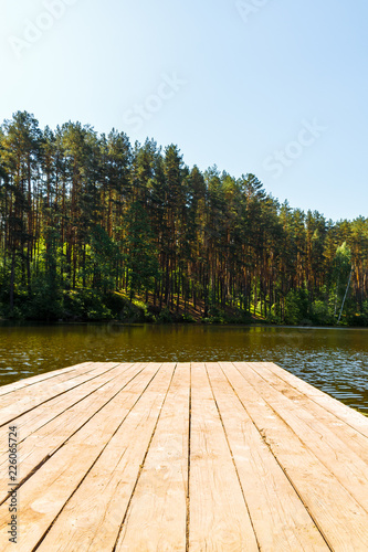 Wooden pier on the river bank in tall green pines under a blue sky.