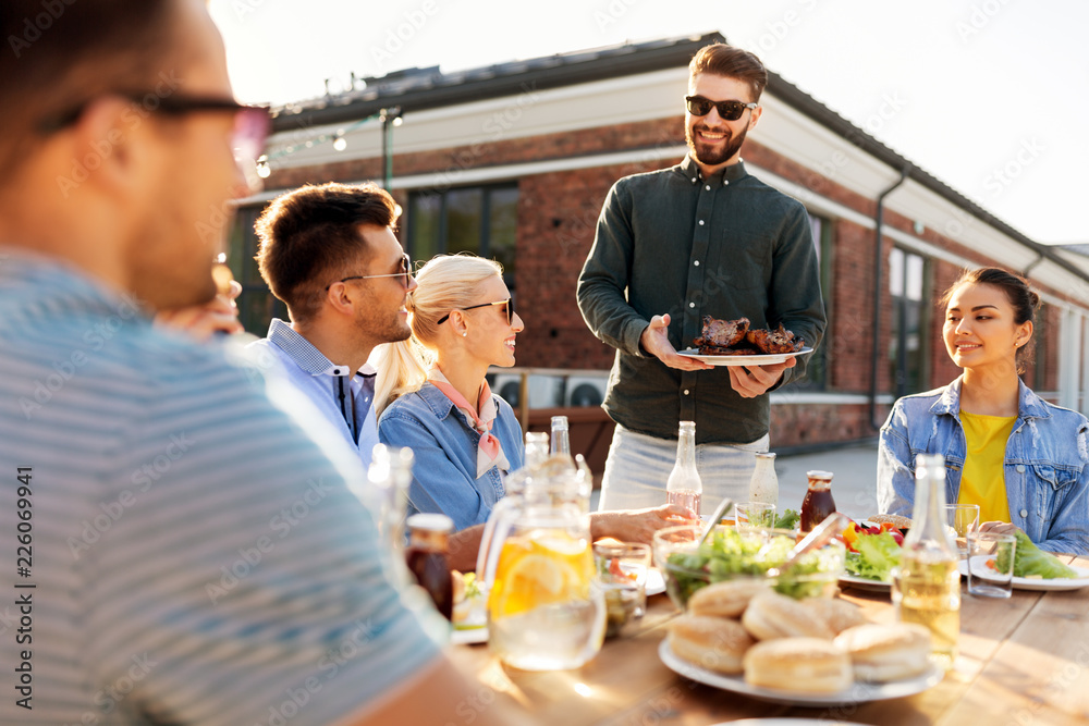 leisure and people concept - happy party host offering meat to his friends at barbecue party on rooftop in summer leisure and people concept - happy party host offering meat to his friends at barbecue party on rooftop in summer