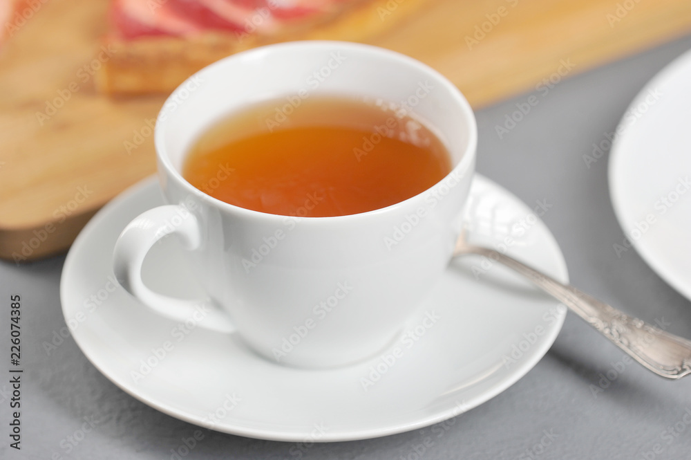 White cup with tea. On a saucer a teaspoon. In the background is an apple pie. Light background. Close-up. White cup with tea. On a saucer a teaspoon. In the background is an apple pie. Light background. Close-up.