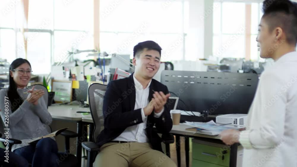 Medium shot of group of young Asian people sitting in chairs in office and listening to female boss make announcement, then clapping hands and smiling Medium shot of group of young Asian people sitting in chairs in office and listening to female boss make announcement, then clapping hands and smiling