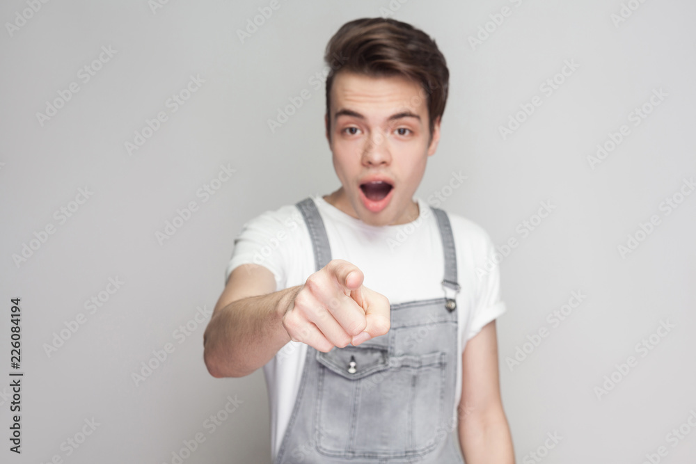 Amazed young brunette man in casual style with t-shirt and denim overalls standing, looking and pointing at camera with surprised unbelievable face. indoor studio shot, isolated on gray background. Amazed young brunette man in casual style with t-shirt and denim overalls standing, looking and pointing at camera with surprised unbelievable face. indoor studio shot, isolated on gray background.