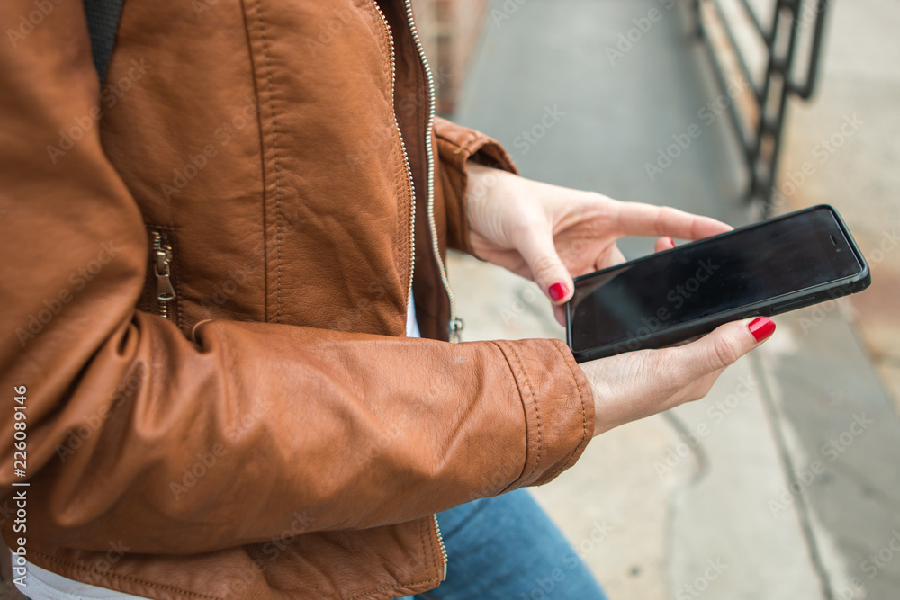 Close up of woman in brown jacket using smartphone online messaging in the street, female hands typing text message via cellphone, social networking concept