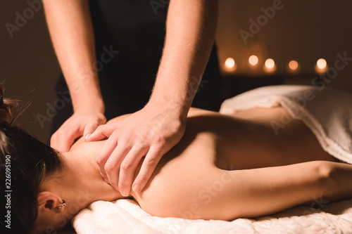 Men s hands make a therapeutic neck massage for a girl lying on a massage couch in a massage spa with dark lighting. Close-up. Dark Key