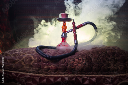 Hookah hot coals on shisha bowl making clouds of steam at Arabian interior. Oriental ornament on the carpet. Stylish oriental shisha in dark with backlight. For Shisha advertisement. Selective focus photo