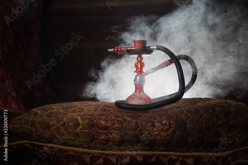 Hookah hot coals on shisha bowl making clouds of steam at Arabian interior. Oriental ornament on the carpet. Stylish oriental shisha in dark with backlight. For Shisha advertisement. Selective focus photo