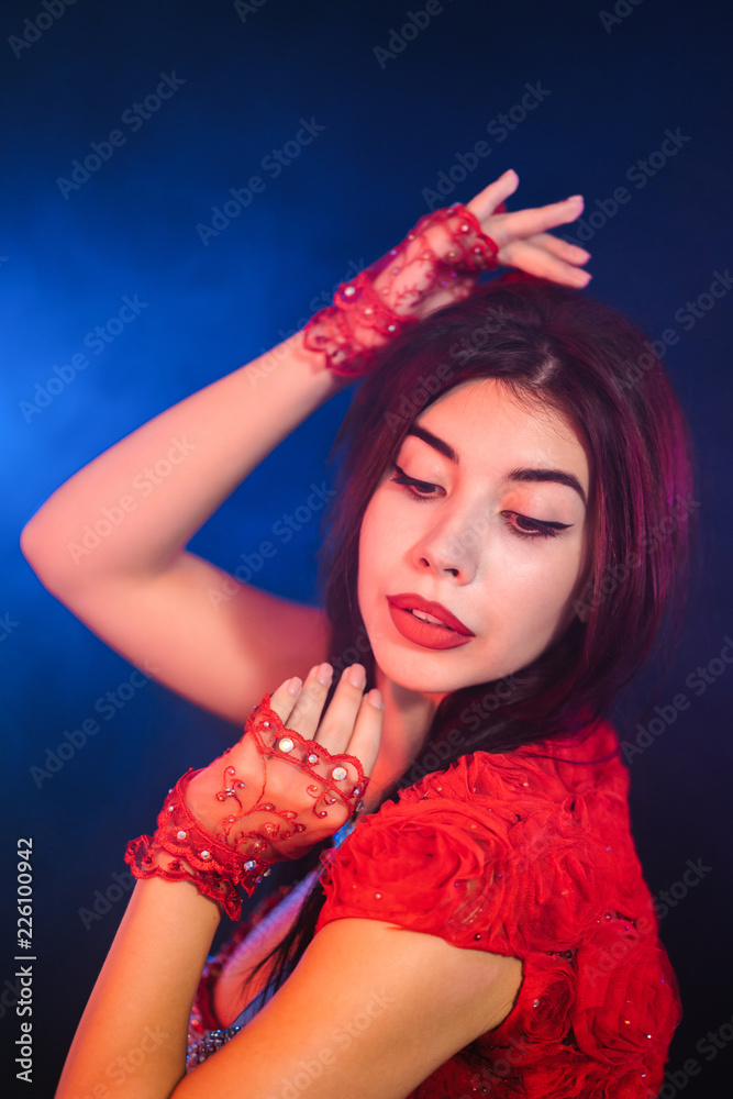 Fototapeta premium Tempting sexy traditional oriental belly dancer girl dancing on purple neon smoke background. Woman in exotic red costume sexually moves her semi-nude body.