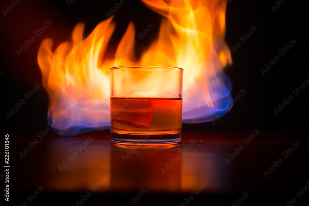 Whiskey in fire concept. Glass of whiskey and ice on wooden surface with color light and fog on background. Close up. Whiskey in fire concept. Glass of whiskey and ice on wooden surface with color light and fog on background. Close up.