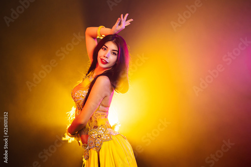 Seductive sexy traditional oriental belly dancer girl dancing on yellow neon smoke background. Woman in exotic costume with feathers sexually moves her semi-nude body.