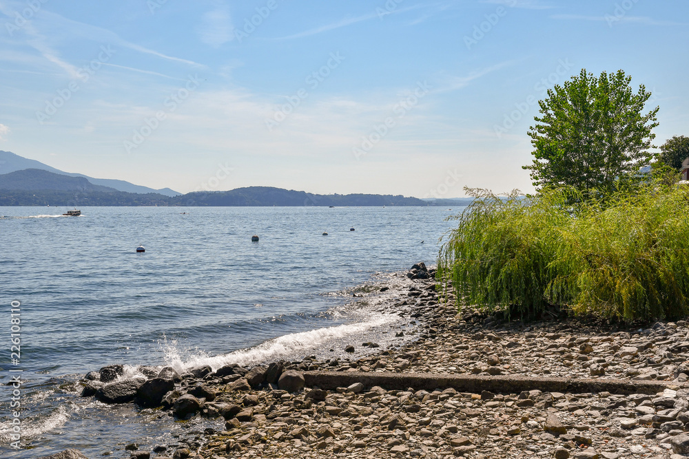 Spiaggia di sassi con veduta del lago in estate con cielo blu e montagne sullo sfondo, Isola dei Pescatori, Lago Maggiore, Piemonte, Italia