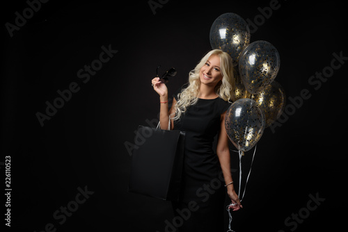 Elegant blonde woman wears sunglasses and black dress holding black shopping bags and festive balloons, black friday concept