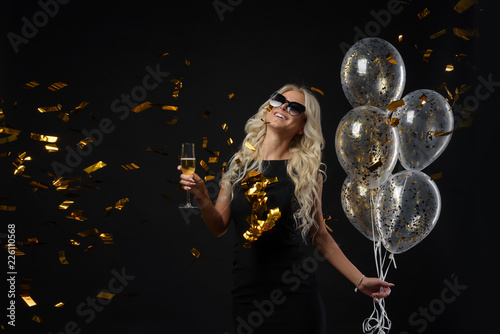 Brightfull expressions of happy emotions of  amazing blonde girl celebrating party on black background. Luxury black dresses, smiling, a glass of champagne, golden tinsels,  balloons, long curly hair