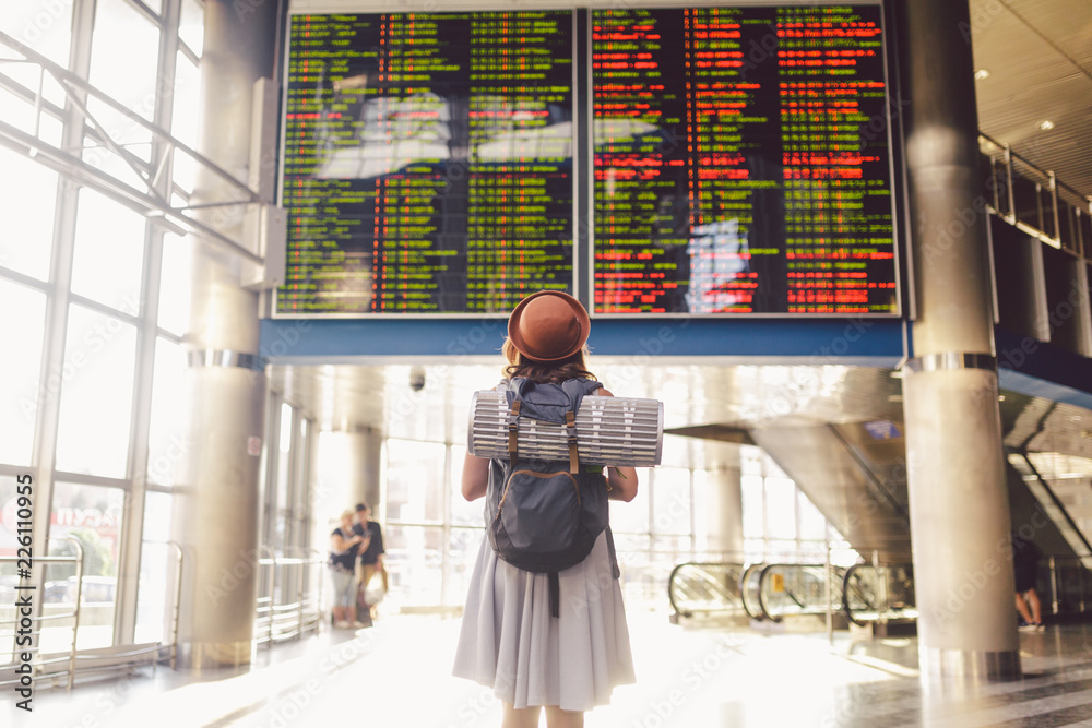 Theme travel public transport. young woman standing with back in dress and hat behind backpack and camping equipment for sleeping, insulating mat looks schedule on scoreboard airport station