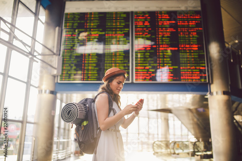 Theme travel and tranosport. Beautiful young caucasian woman in dress and backpack standing inside train station or terminal looking at a schedule holding a red phone, uses communication technology photo