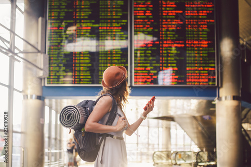Theme travel and tranosport. Beautiful young caucasian woman in dress and backpack standing inside train station or terminal looking at a schedule holding a red phone, uses communication technology photo