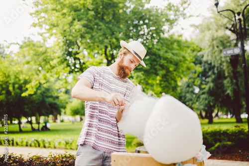 photo theme small business cooking sweets. A young man with a beard of a Caucasian trader in the hat the owner of the outlet makes candy floss, fairy floss or Cotton candy in the summer park