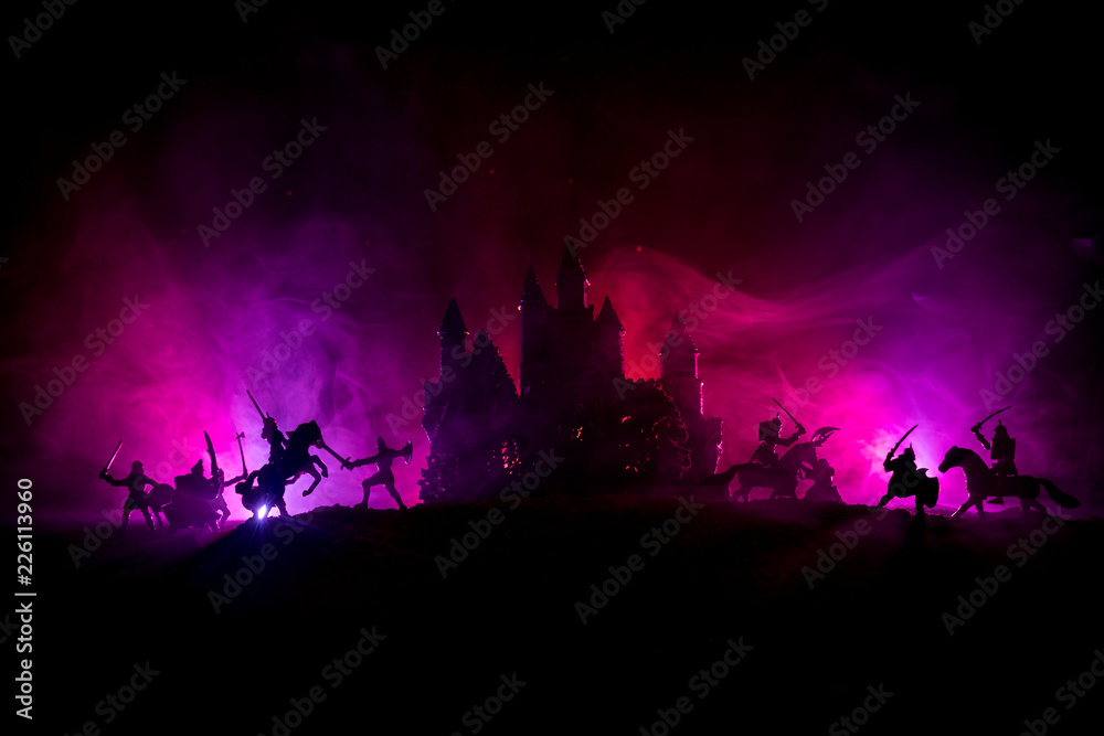 Medieval battle scene with cavalry and infantry. Silhouettes of figures as separate objects, fight between warriors on dark toned foggy background with medieval castle. Night scene. Selective focus Medieval battle scene with cavalry and infantry. Silhouettes of figures as separate objects, fight between warriors on dark toned foggy background with medieval castle. Night scene. Selective focus