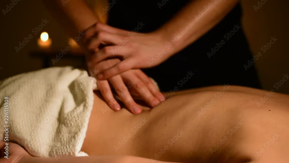 Close-up spa massage woman's shoulders and back. Male hands do massage to a woman in a dark room with candles in the background Close-up spa massage woman's shoulders and back. Male hands do massage to a woman in a dark room with candles in the background