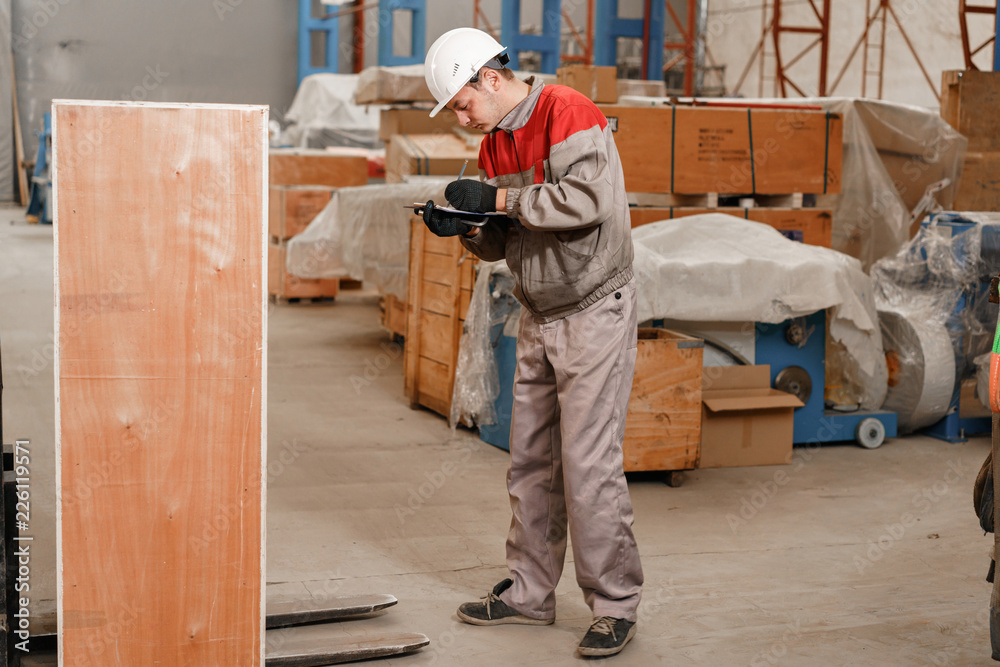 the storekeeper marks the arrival on the tablet. man driving a forklift through a warehouse in a factory. driver in uniform and protective helmet. the concept of logistics and storage the storekeeper marks the arrival on the tablet. man driving a forklift through a warehouse in a factory. driver in uniform and protective helmet. the concept of logistics and storage