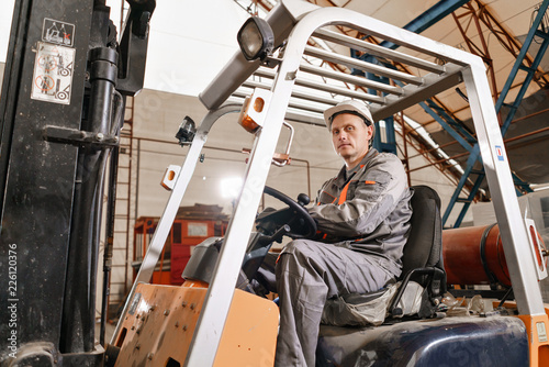 man driving a forklift through a warehouse in a factory. driver in uniform and protective helmet. the concept of logistics and storage