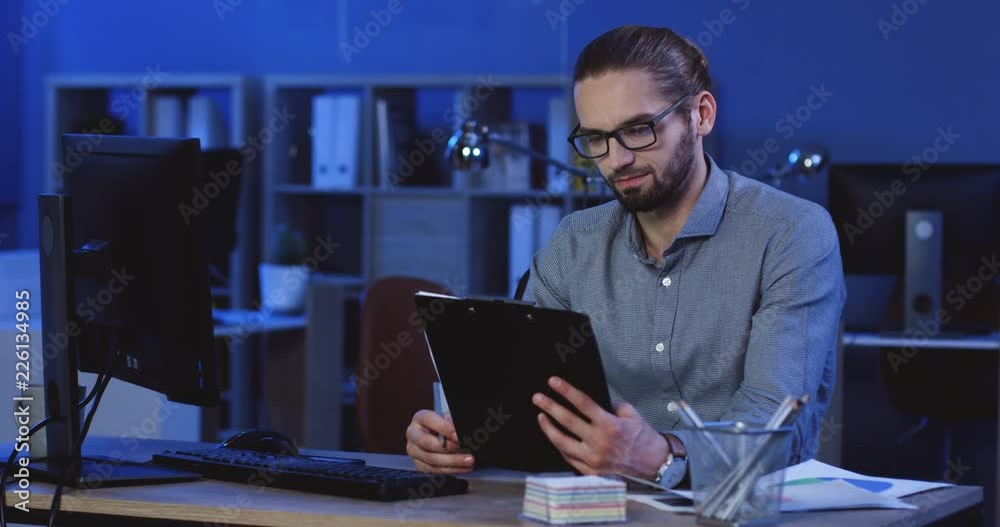 Caucasian handsome young businessman reading some documents or contract and doing notes on it in his cabinet late in the evening.