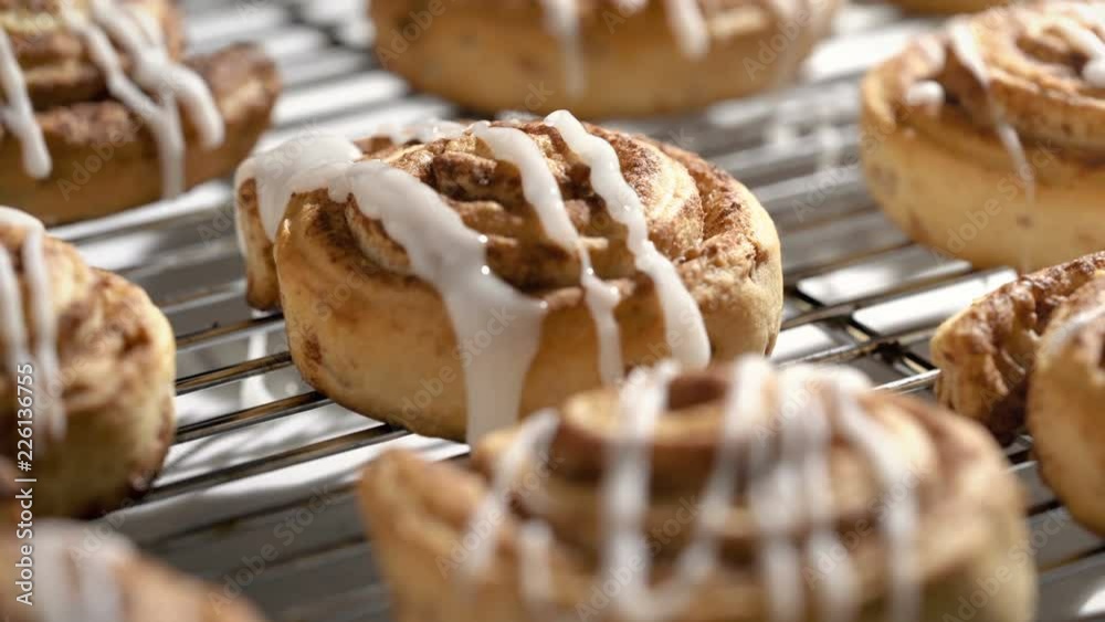 Cinnamon bun on cooling rack with white icing being dripped over the fresh baked pastries. 