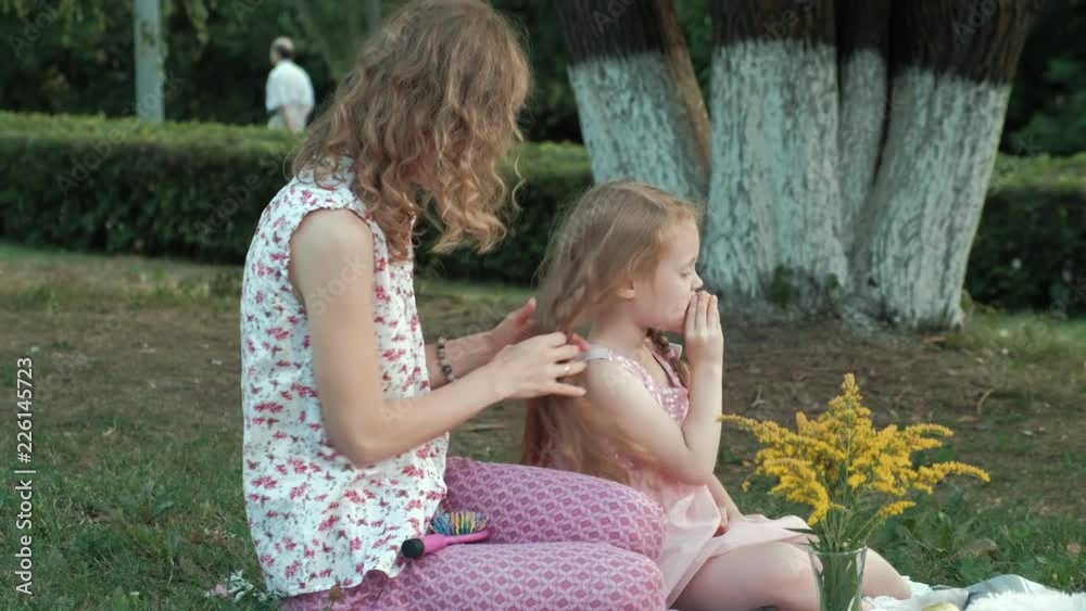A happy mother combs her daughter's hair. Family in a city park on a picnic on a warm evening at sunset. A happy mother combs her daughter's hair. Family in a city park on a picnic on a warm evening at sunset.