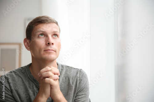 Man with hands clasped together for prayer near window. Space for text
