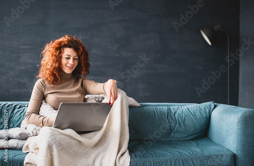 Redhead young woman with curly hair sitting on the couch, looking at the tablet screen