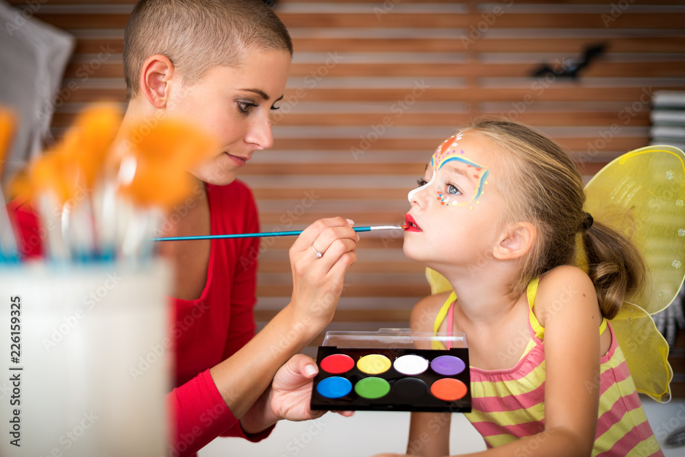 Fototapeta premium Young mother painting daughters face for Halloween party. Halloween or carnival family lifestyle background. Face painting and dressing up.