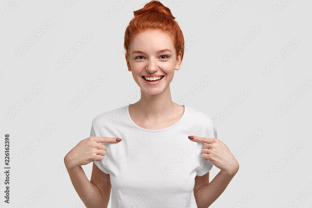 Horizontal shot of pleasant looking ginger girlfriend points at copy space of her clothes, shows place for your promotional content, smiles positively, isolated on white background. Clothing concept Horizontal shot of pleasant looking ginger girlfriend points at copy space of her clothes, shows place for your promotional content, smiles positively, isolated on white background. Clothing concept