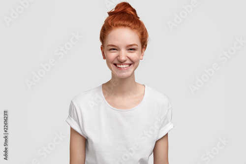 Adorable freckled young lady or teenager smiles joyfully at camera, has red hair combed in knot, dressed at casual t shirt in one tone with background, thinks about good things. Emotions concept
