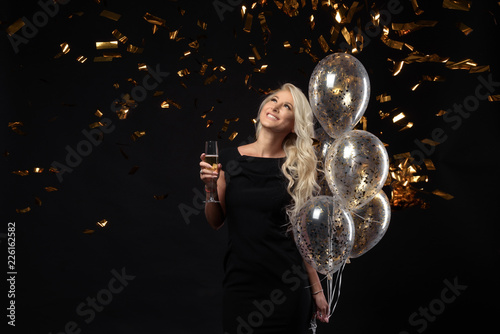 Brightfull expressions of happy emotions of  amazing blonde girl celebrating party on black background. Luxury black dresses, smiling, a glass of champagne, golden tinsels,  balloons, long curly hair