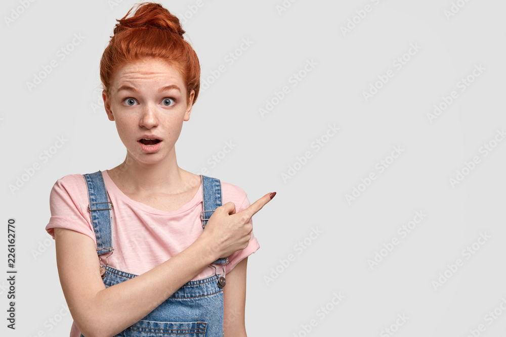 Photo of astonished red haired freckled girl indicates with index finger at upper right corner, keeps jaw dropped from amazement, poses against white background, copy space for your advertisement Photo of astonished red haired freckled girl indicates with index finger at upper right corner, keeps jaw dropped from amazement, poses against white background, copy space for your advertisement