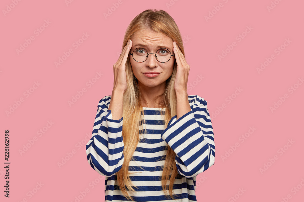 Stressful overworked busy female with displeased frustrated expression keeps hands on temples, dressed in casual striped sweater, isolated over pink background, has terrible headache. Omg, what to do? Stressful overworked busy female with displeased frustrated expression keeps hands on temples, dressed in casual striped sweater, isolated over pink background, has terrible headache. Omg, what to do?