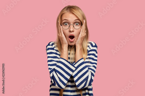 Photo of stupefied blonde pretty woman stares with bugged eyes at camera, opens mouth widely, dressed in casual striped sweater, stands against pink background. People, surprisement and disbelief photo