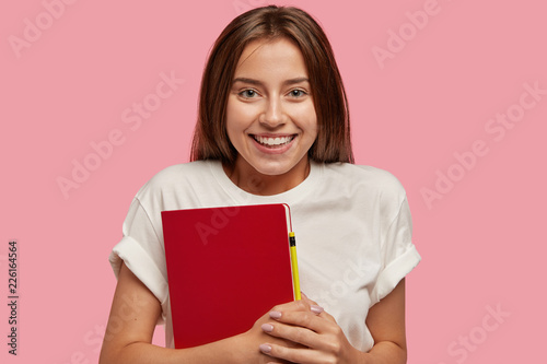 Cheerful teenage girl has toothy smile, healthy skin, long dark hair, carries red notepad with pencil, makes notes, dressed in casual white t shirt, satisfied with successful day, models indoor alone