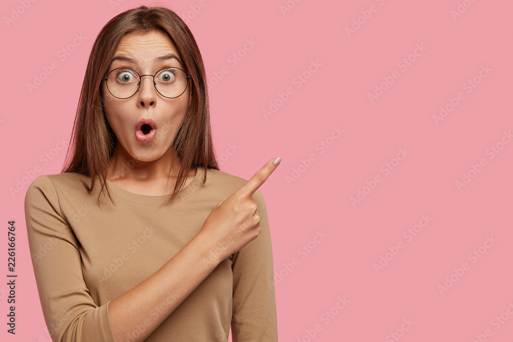 Photo of stupefiied Caucasian girl stares at camera, keeps jaw dropped, points with index finger at upper right corner, shows free space for your promotional content. Omg, look at this thing Photo of stupefiied Caucasian girl stares at camera, keeps jaw dropped, points with index finger at upper right corner, shows free space for your promotional content. Omg, look at this thing