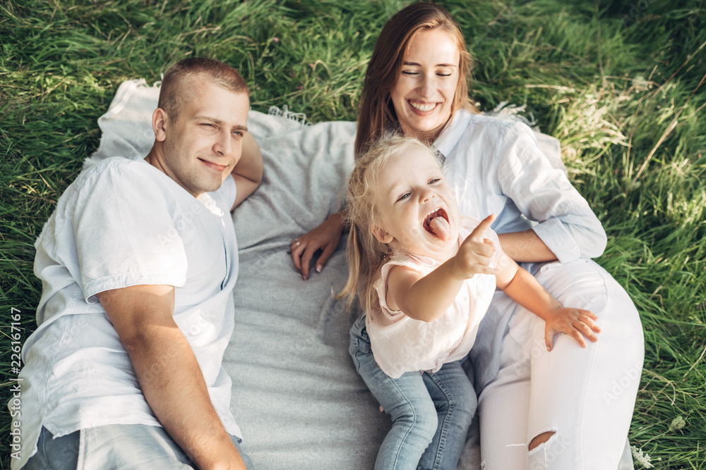 Young Adult Couple with Their Little Daughter Having Fun in the Park Outside the City, Family Weekend Picnic Concept, Three People Enjoying Summer Time Young Adult Couple with Their Little Daughter Having Fun in the Park Outside the City, Family Weekend Picnic Concept, Three People Enjoying Summer Time