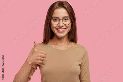 Portrait of happy teenager keeps thumb raised, being in good mood, shows her agreement, poses over pink background, says: Nice joke! Young woman shows like gesture, satisfied with something.