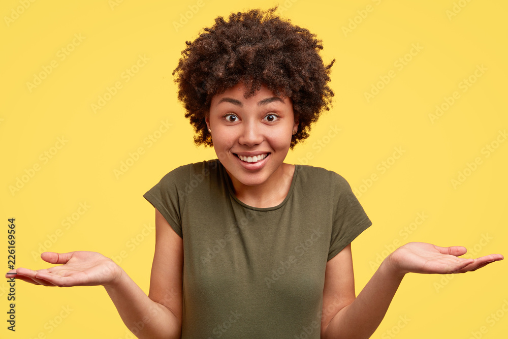 Uncertain clueless cheerful dark skinned lady in casual military t shirt, shruggs shoulders with hesitation, has charming smile, poses against yellow background. Ethinicty and body language concept