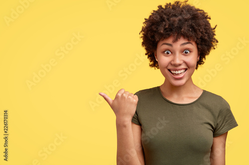 Lovely woman with overjoyed expression points aside, shows free space for your advertisement, dressed in casual military t shirt, cant believe in so high discounts, recommends to visit this shop