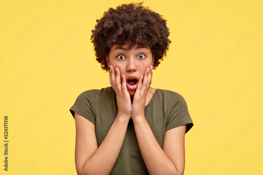 Gosh, something awful happening. Impressed amazed woman with Afro hair dropps jaw, keeps both hands on cheeks, dressed in casual t shirt, poses against yellow background. Surprisement and shock Gosh, something awful happening. Impressed amazed woman with Afro hair dropps jaw, keeps both hands on cheeks, dressed in casual t shirt, poses against yellow background. Surprisement and shock