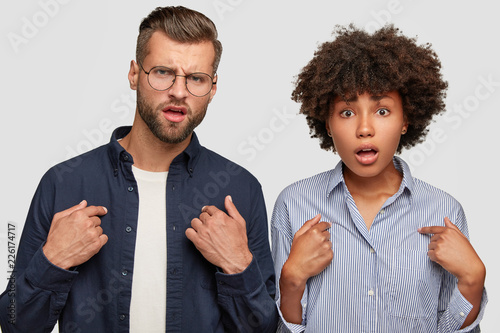 Horizontal shot of indignant young female and male of different races, point at themselves, ask something, stand next to each other, being shocked and displeased. Why should we prepare this task?