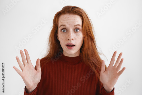 Oh my God. Isolated shot of surprised beautiful young red haired female in turtleneck raising eyebrows and keeping mouth opened, gesturing emotionally, listening to incredible shocking news