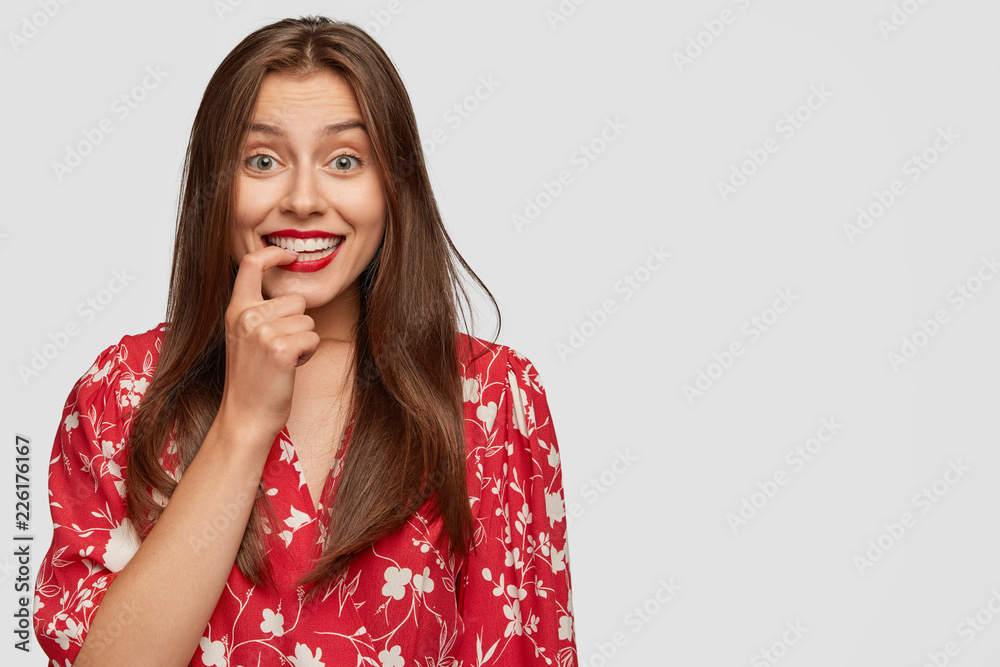 Happy unexperienced businesswoman smiles positively, keeps finger on teeth, thinks about fulfillment of future plan, has interesting idea, stands against white background with blank copy space Happy unexperienced businesswoman smiles positively, keeps finger on teeth, thinks about fulfillment of future plan, has interesting idea, stands against white background with blank copy space