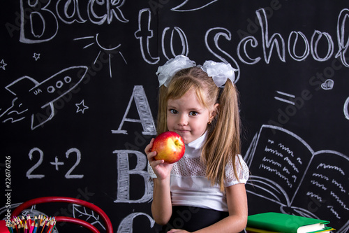 Greedy schoolgirl sitting on the desk with books, school supplies, yholding a red apple in the right hand