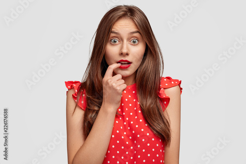 Omg, why it happens with me? Good looking brunette woman has dark hair, stares with bugged eyes, bites finger, dressed in polka dot blouse, cant believe in shocking news, isolated on white wall